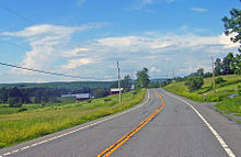 A two-lane highway in a sparsely developed and hilly area with two farm buildings nearby. Large mountain ridges are visible in the distance.