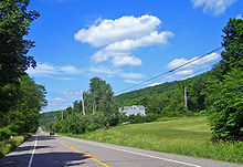 A two-lane highway lined with telephone poles passes through a rural, forested area. In the background is a tree-covered ridge.