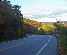 A curving highway descending into the left of the image surrounded by woods, some of which are showing fall color. Mountains loom in the distance on the right.