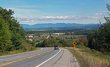 A road curving to the left with a car on it and a sign warning of a steep grade. It is descending toward a lower area with some buildings and another stretch of road. In the distance is a line of mountains.