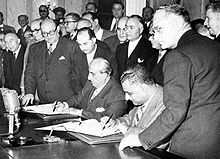 Men in jackets standing, while two are sitting next to each other, each holding a pen and writing something on paper.