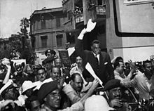 Men wearing different clothes are marching while in their center there is a man standing on a car, while holding his hand up with a towel. There are men in military uniform, some in Jellabiyas and others in jackets.