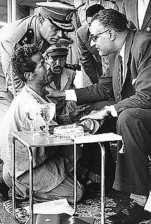 A man on his knees looking up to a man sitting and holding his hand and wearing sun glasses, has his right hand on his shoulder and is talking to him. In the background there are men in military uniform all looking on the kneeling man.
