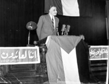 A man holding his hands back, while standing on a stage with three microphones in front of him and a lamp on his side. A flag with three different color stripes and a triangle at the top is hung in front of him at the stage.