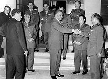 A group of men most in military uniform, but some wearing jackets. In the center a man in a gray jacket is shaking hands with another military figure, while most men are looking at them and smiling.