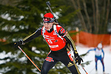 A woman in predominantly black winter sportswear, wearing a red cap and a bright red jersey with the number 13, is pictured in a forested area covered with snow. She leans slightly forward and holds ski poles in her hands. There is a green conifer tree behind her and a second person can be seen in the background out of focus on the right.