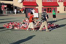 A makeshift memorial outside of a stadium. In the center of the picture are baseball caps, baseballs, and flowers underneath a picture as two people walk by, glancing at the memorial.