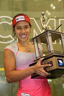 A young woman in a white-and-pink shirt and a red headband hoists a large squarish trophy, which has four pillars and a figurine at top.