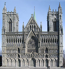 A Romanesque church with Gothic pointed arches. Two rectangular towers separated by a pitched roof entrance hall. The ornate facade is heavily articulated with repeated arches, most of them framing statues.