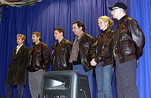 Six actors, all but one wearing a leather jacket, are photographed on a stage with a blue curtain as a backdrop.
