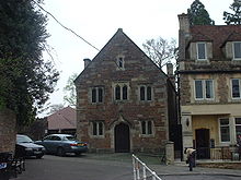 Stone building with arched doorway on ground floor. Three windows on first floor and single window below roof level. To the left are trees and cars, to the right another building.