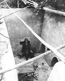 A soldier sitting on a chair in a large exposed underground room. To the right are two large holes in the wall while the room is littered with other furniture, including a makeshift table.