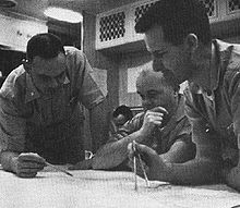 Three naval officer plotting the submerged course of the nuclear submarine Triton, with Captain Beach seated before chart in the center, with Lt. Commander Will M. Adams standing at right holding a pair of dividers abd Lt. Commander Robert W. Bulmer standign at the left holding a pencil, and an unidentified individual seated in the background.