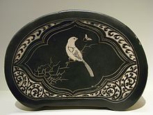 An oval shaped pillow with flat sides. It is made of black cloth with an image of a white bird sitting on a branch stitched into it.