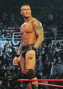 A caucasian male with closely cropped dair hair stands on the turnbuckles of a wrestling ring with red ropes. He has 'sleeve' tattoos covering both his arms, black wrist tape around both wrists, and is wearing short black wrestling tights and black kneepads.