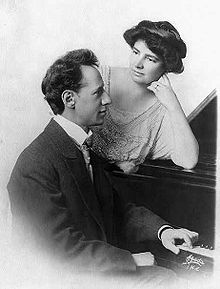 Ossip and Clara (in her forties) at a piano showing the upper half of their bodies.  Ossip sits at the piano looks to the right with one hand resting on the keyboard.  Clara is standing and leans her elbow on the top of the piano and looks at Ossip. Her hair is dark short and styled in waves.