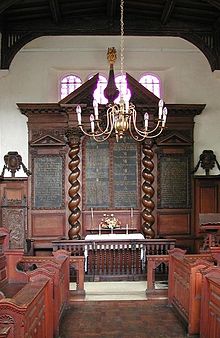 The interior of a chapel showing an elaborate wooden reredos with curling columns and inscribed panels, in front of which is a candelabrum and benches