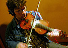 Seated man playing a fiddle in a studio, wearing earphones.