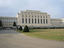 A drive leads past a manicured lawn to large white rectangular building with a columns on it facade. Two wings of the building are set back from the middle section.