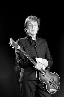 Black-and-white image of McCartney, in his sixties, holding an electric bass. He wears a black buttoned-up suit jacket with black pants.