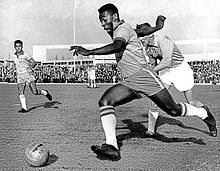 A dark-skinned young man in soccer attire is pictured in mid-stride as he sprints with the ball past an opposing player, who looks tired and has given up attempting to chase him.