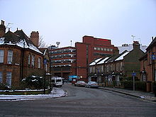 A tall red brick building towers over streets of two-storey houses. The roofs of the houses and the surrounding pavements are covered in snow