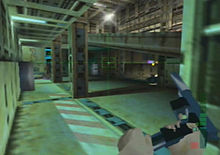 A metallic room with colums and computers located in the farthest side. A hand holding and reloading a gun is seen on the bottom right corner. A crosshair and graphics symbols representing ammunition are also visible.