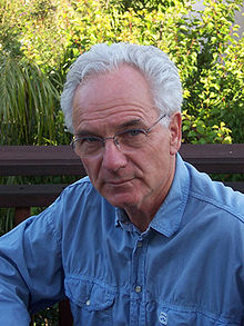Peter Duesberg, a white male with grey hair and a blue shirt, sitting in front of a stand of plants