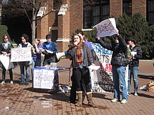 A college-aged female in jacket and scarf holds the microphone attachment of a bullhorn while other students hold protest signs behind her. Two with large red X's over the words read "Free Speech" and "Access".