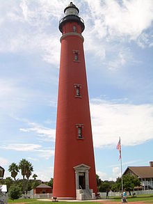 A tall red lighthouse, seen from the point of view of someone on the ground, is seen framed by a blue sky, with palm trees and a flagstaff in the background.
