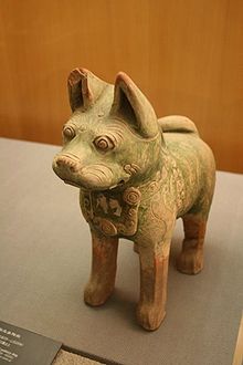 A green-glazed ceramic statuette of a dog with pointy ears and curly tail, standing upright on all fours with eyes open