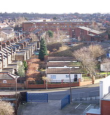 A strip of grass recedes in a straight line into the distance towards a large yellow brick building. Three single storey huts stand on the grass