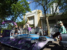 Young woman standing on a purple float, behind a sign that says "Princess Kay of the Milky Way". Float is passing the Horticulture building. Fairchild, the fair's mascot, is at right.