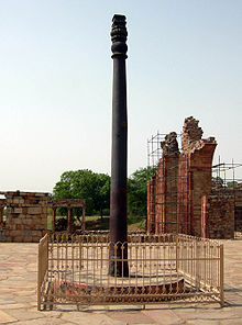 An pillar, slightly fluted, with some ornamentation at its top. It is black, slightly weathered to a dark brown near the base. It is around 7 meters (22 feet) tall. It stands upon a raised circular base of stone, and is surrounded by a short, square fence.