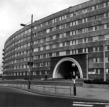 A black-and-white photograph of part of a monumental seven-storey curved-fronted block of flats made of poured and pre-cast concrete with a prominent two-storey semicircular entrance arch. In the foreground is a pedestrian crossing with a Belisha Beacon.