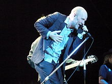 A middle-aged Caucasian man in a blue pin stripe suit with a shaved head sings into a microphone and smiles.
