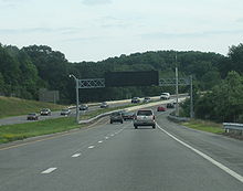 Ground-level view of a four-lane divided highway with a wide grassy median separating the opposing lanes of traffic; in the distance the road makes a sharp curve. A black electronic sign without lettering is visible above the highway.