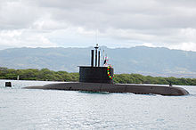 A submarine travelling on the water's surface near to shore, with trees and mountains in the background.