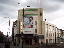 A large cream-coloured and tiled building stands at the intersection of two roads. Dark grey clouds dominate an overcast sky. Two flags are flying from the fascia of the building, which is covered mostly by a large advertising hoarding.