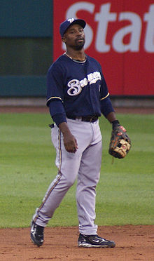 A man wearing gray pants, a navy blue jersey with "Brewers" written in white, a navy blue cap bearing a white "M", and a baseball glove stands on a baseball field