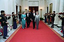 Long shot of two men flanked by two women walking down read carpet, as military band plays on either side