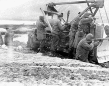 Seven men with rifles firing from behind a bulldozer, in a barren landscape with river and mountains in distance