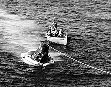 A conical black spacecraft, with its base surrounded by inflated bags, floating in the water; a rope runs from the top of the spacecraft to the edge of the photograph. A small boat, with a group of men in it is just behind the spacecraft.