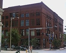 A large four-story red brick building with many windows. The building is on a street corner with the front and one of the sides visible.