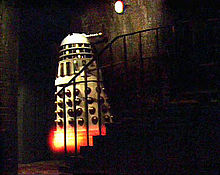 In a dark basement, a white Dalek (see previous description) appears to levitate up a small staircase of approximately seven stairs. The body of the Dalek is white, with shiny gold vertical slats and gold balls on its lower half. There is an orange-yellow glow at the Dalek's base.