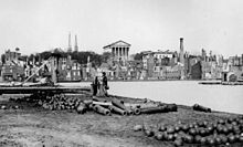 Wide view of a cityscape with evident destruction. Unused cannons and cannonballs litter the foreground, while a large Neoclassical building stands intact in the rear center.