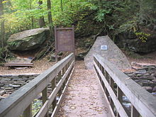 View from a wooden footbridge with handrails over a rocky creek in a green forest. At the end of the bridge are a large boulder with a bench below it to the left, a trail sign labeled "Waters Meet" and "The Falls Trail" above a map of the trail in the center, and a natural stone monument with a metal plaque to the right.