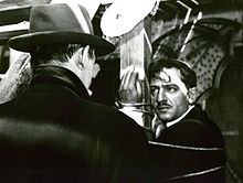 A screen capture from the film, showing a distraught Cesar tied up to a pole staring at Tony le Stephanoi who has his back to the camera.