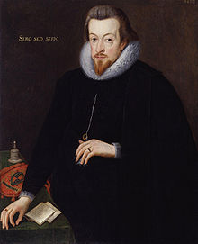 A portrait of Robert Cecil, who is standing at a table wearing black robes.  He has neck length brown hair, and a pointed goatee.  He has gold lettering behind him, which reads "sero, sed serio."
