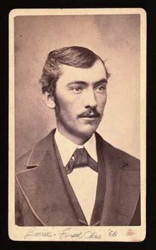 A mustachioed man in a dark jacket with a broad collar, white shirt, and dark tie.